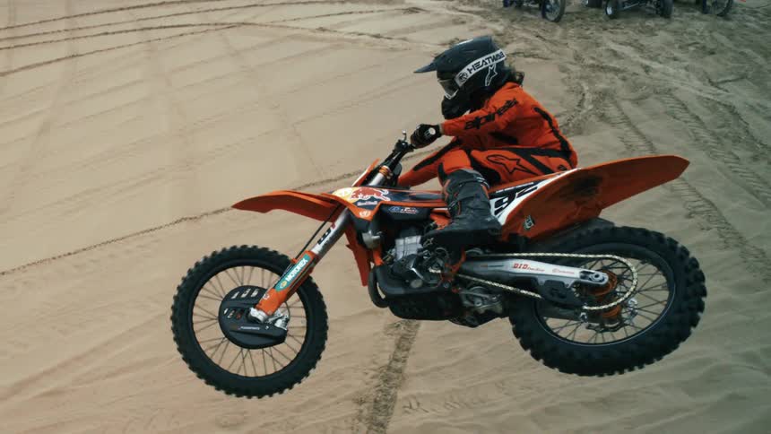 A motocross rider is captured mid-air in slow motion during a spectacular jump over sand dunes