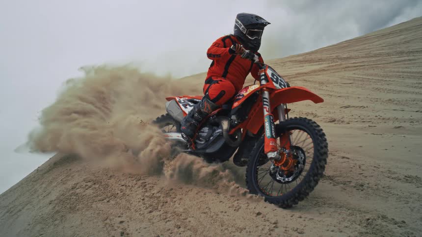 A dirt bike rider in an orange suit expertly carves a turn on a steep sand dune, spraying sand in dramatic slow motion
