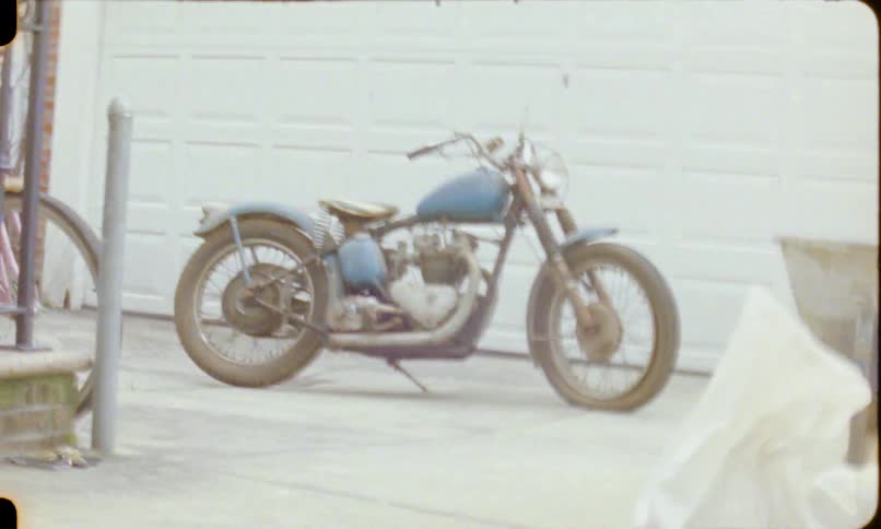 Vintage 8mm film captures a classic blue motorcycle parked in a driveway before the footage becomes distorted by a light leak