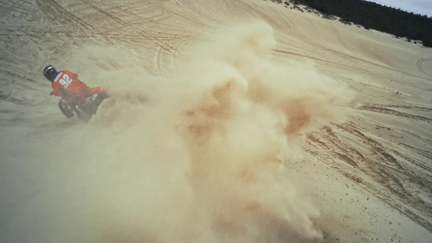 An epic high-angle slow-motion shot captures a motocross rider kicking up a massive cloud of sand while turning on a dune