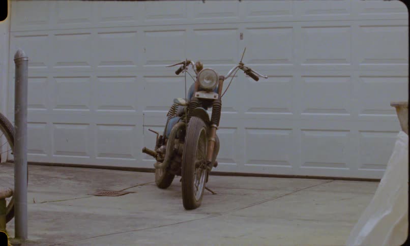 A vintage blue motorcycle parked on a concrete driveway in front of a white garage door, captured on retro film