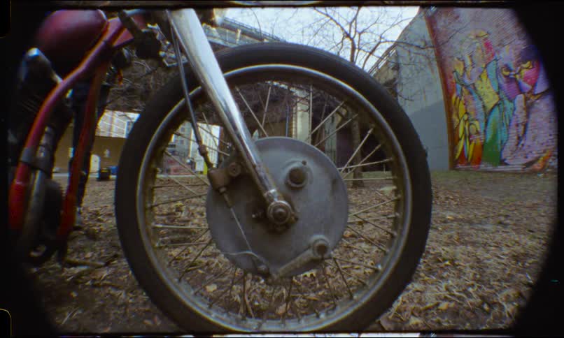 A stylized vintage film close-up of a classic motorcycle's front wheel in a gritty urban lot with a colorful mural in the background