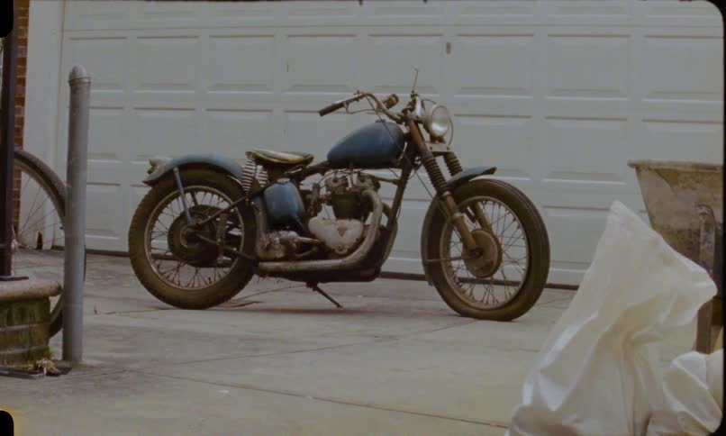 A still shot of a classic blue motorcycle parked on a concrete driveway in front of a residential garage door, captured on vintage film