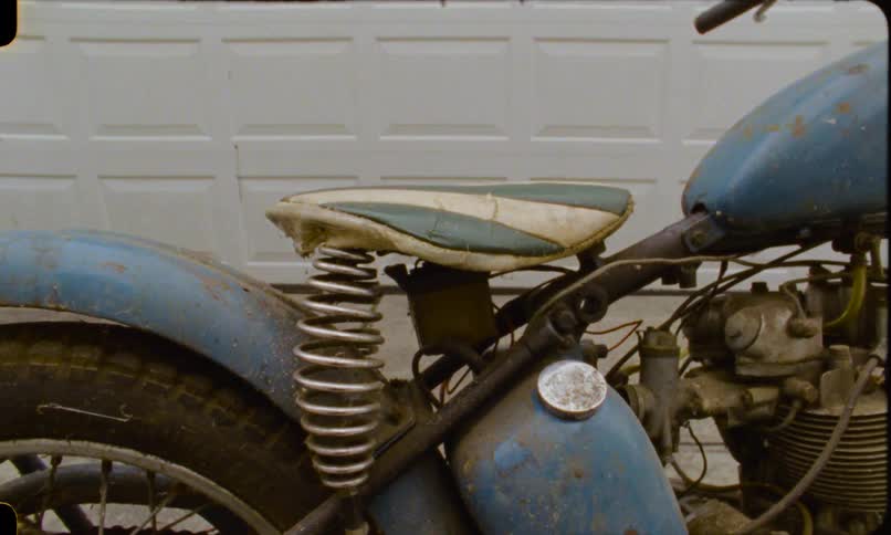 A gritty close-up shot reveals the details of a rusted vintage blue motorcycle parked in front of a garage door