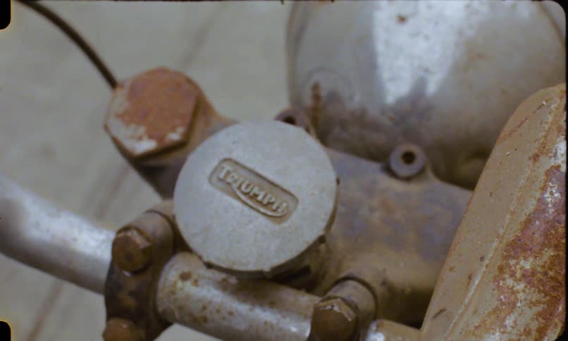 A vintage, grainy film shot shows a close-up of the rusty handlebars and logo cap of an old Triumph motorcycle