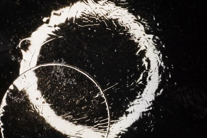 A macro view of a silvery liquid slowly forming intricate circular patterns against a black background