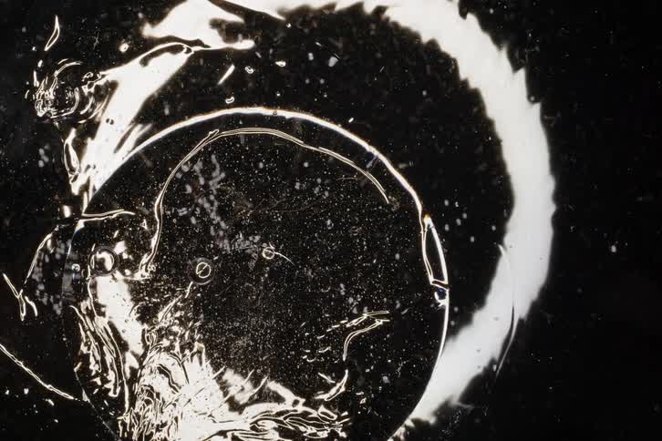 An abstract macro shot of a clear, viscous gel with suspended particles slowly moving and drying on a dark surface