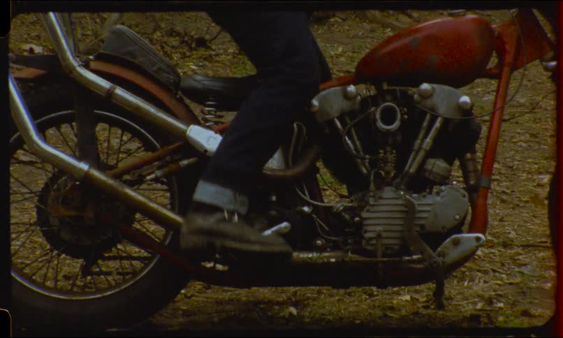 A biker in boots and jeans kick-starts a vintage red chopper, causing a puff of smoke to erupt from the engine
