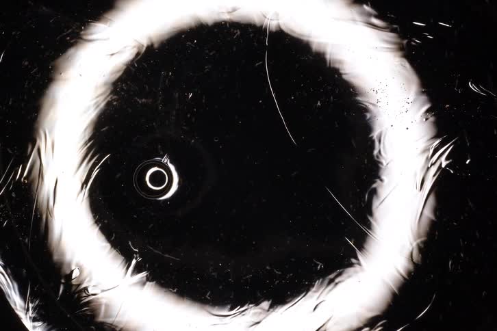 An abstract macro shot of a white glowing ring on a black surface that slowly wrinkles and cracks over time