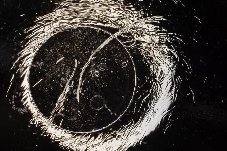 A top-down abstract shot of a clear liquid swirling and creating ripples over circular glass objects against a black background