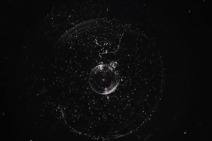 An abstract macro top-down view of a clear bubble slowly shrinking and then popping in a splash against a black background