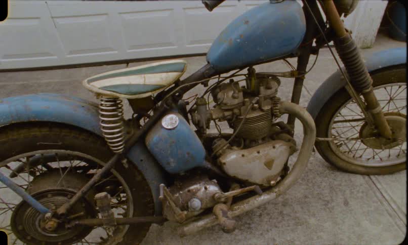 A slow pull-out shot reveals a classic, weathered blue Triumph motorcycle parked on a driveway, shot on vintage film