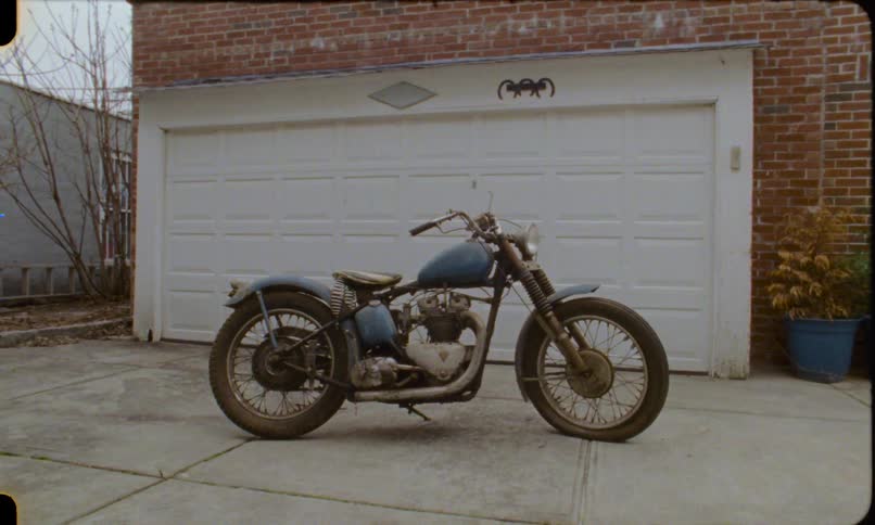 A classic blue motorcycle is parked in a driveway on a cloudy day in this vintage home movie footage