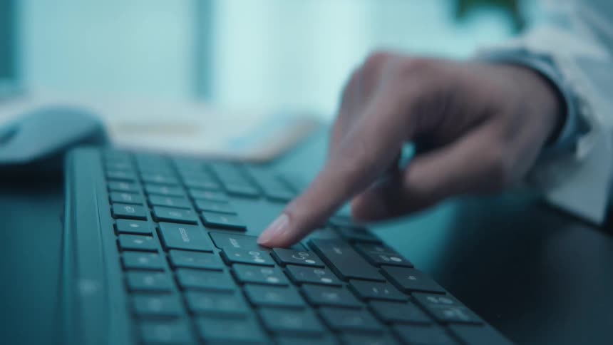 A close-up of a person's hand slowly and deliberately pressing keys on a computer keyboard in a cool, blue-lit room