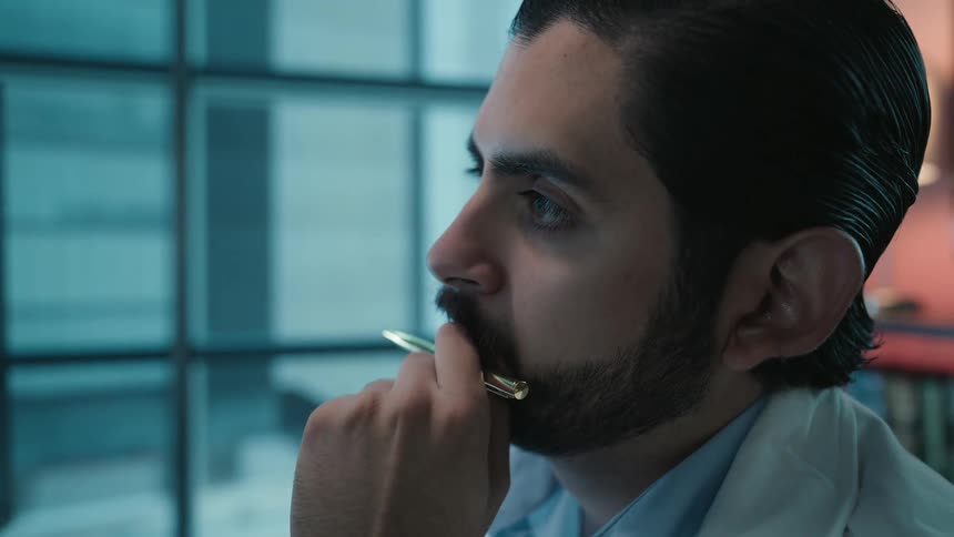 A thoughtful male doctor with a beard sits in his office, carefully analyzing brain scans on a computer monitor and pondering a diagnosis