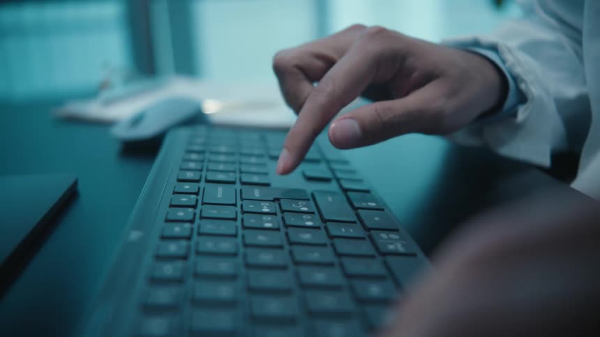 Close up slow motion dolly shot of hands typing on a black keyboard in a modern, blue-lit office setting
