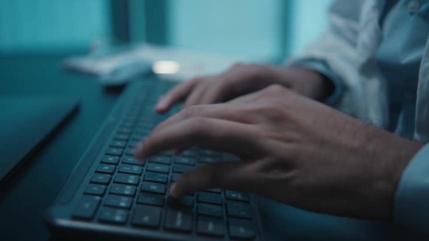 A close-up of a doctor's hands typing on a computer keyboard in a dimly lit office at night