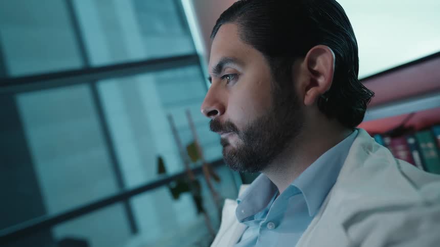 Dolly in on a dedicated male doctor carefully analyzing an X-ray in his modern office late at night