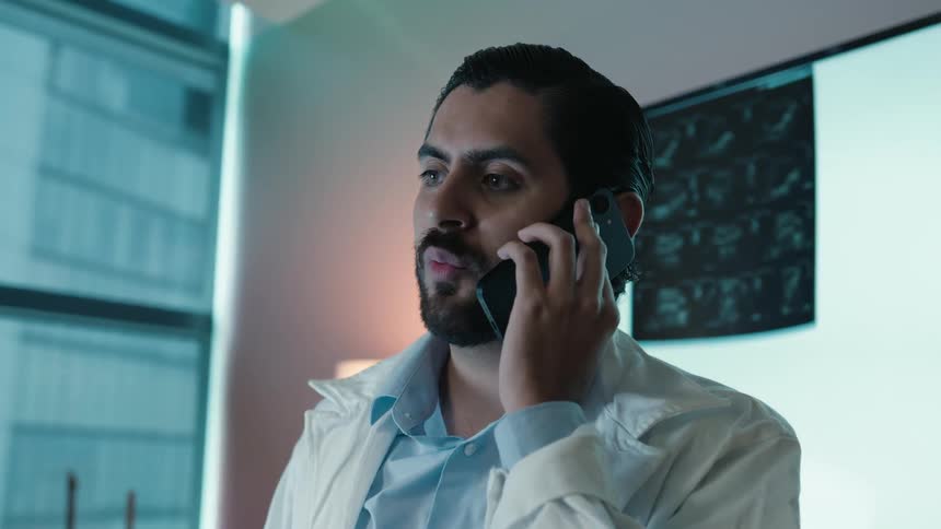 A male doctor in a white coat talks on his smartphone in his office with an x-ray in the background
