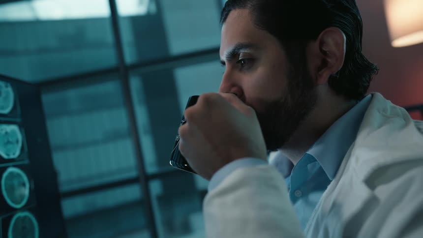 A focused doctor in a white coat sips from his mug while analyzing brain scans on a computer monitor in his dimly lit office