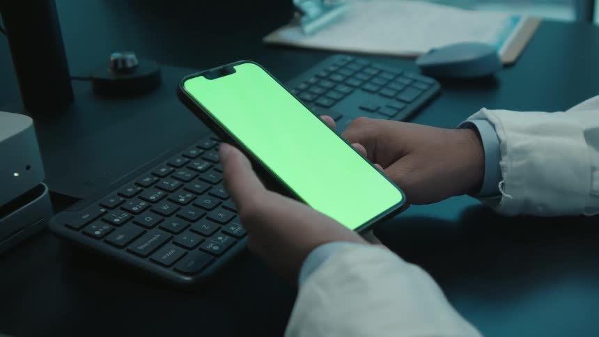 Close up of a doctor's hands holding a smartphone with a green screen while sitting at a desk in a dark office