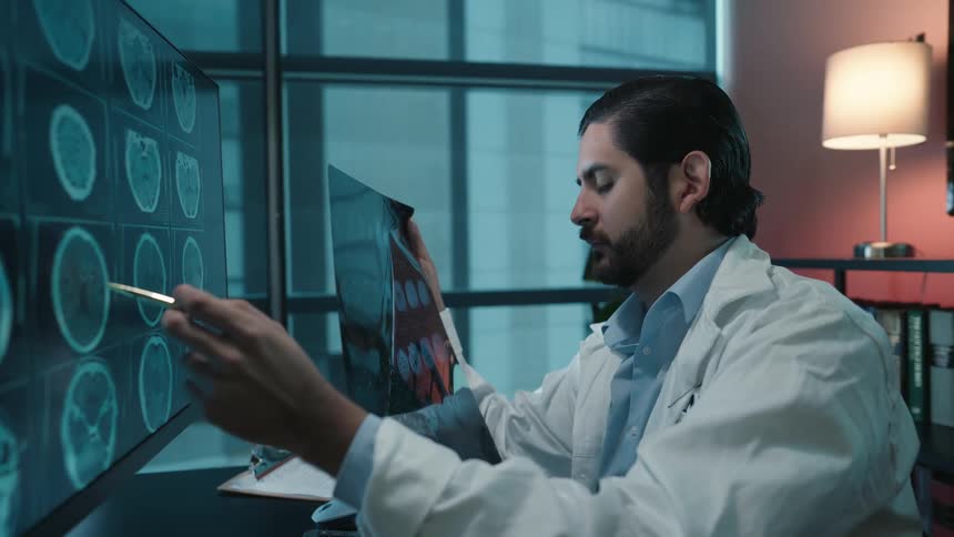 A focused doctor in his office meticulously compares a patient's MRI film to digital brain scans on his computer monitor