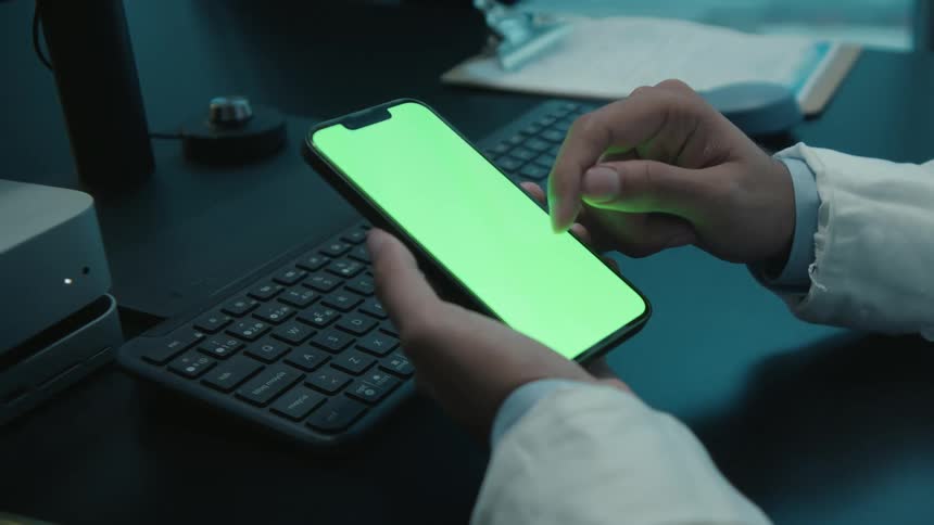 A doctor in a white coat sits at a desk in a dark room and scrolls through a smartphone with a green screen