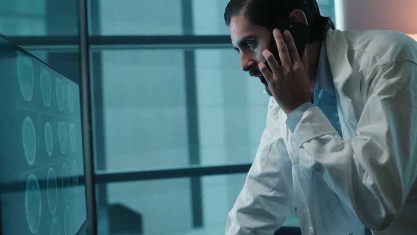 Male doctor in a white coat talks on the phone while closely examining brain scans on a large monitor in a dimly lit office