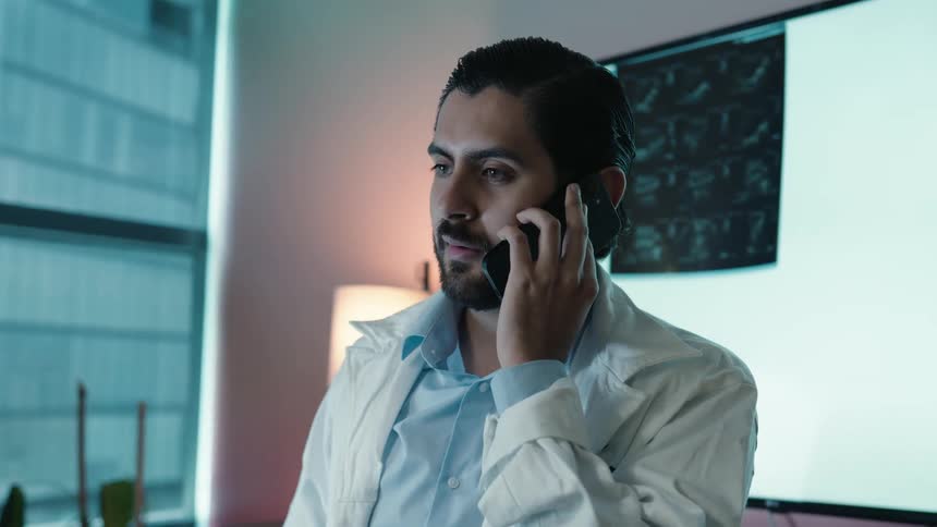 A handsome male doctor in a lab coat discusses a patient's case over the phone in his office at night