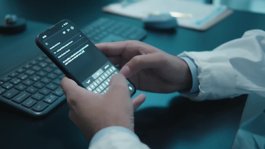 Close-up of a medical professional's hands composing an email on a smartphone at a desk