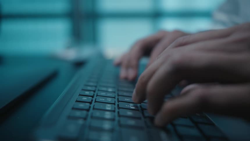 A cinematic side view close-up shows hands typing on a laptop keyboard in a dark, moody office with cool blue lighting