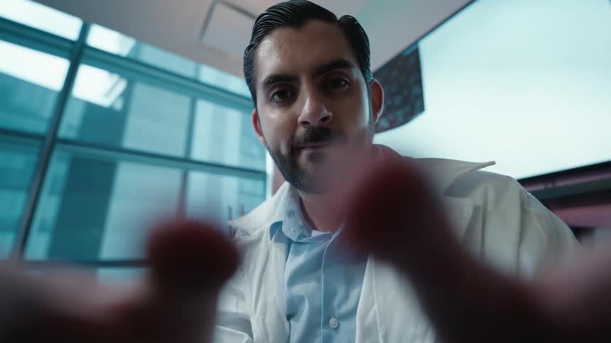 POV of a male doctor in a white lab coat with a focused expression looking down at the camera while his fingers move in the foreground in a looping cinemagraph