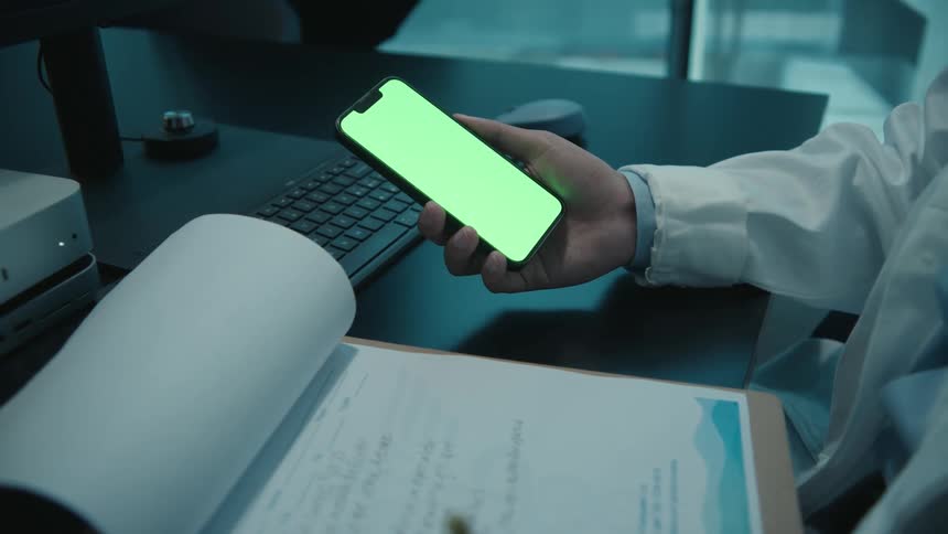 A doctor holds a smartphone with a green screen while taking notes on a clipboard at their desk in a dimly lit office
