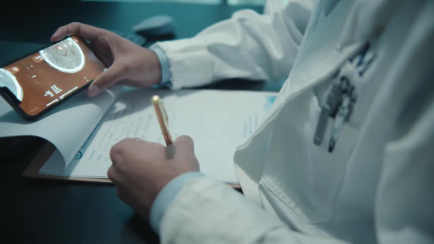 A doctor in a white lab coat analyzes a brain scan on a smartphone while taking notes on a clipboard in a dimly lit office