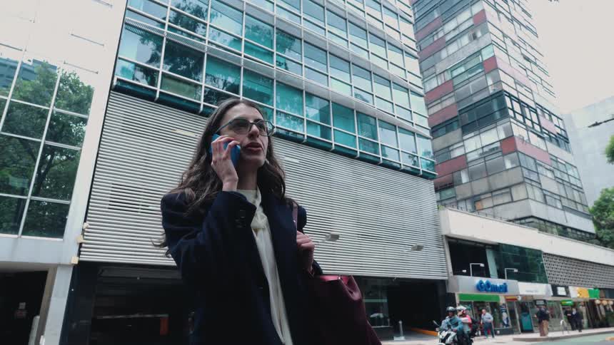 A low-angle shot captures a serious businesswoman talking on her smartphone on a city street with modern buildings looming in the background