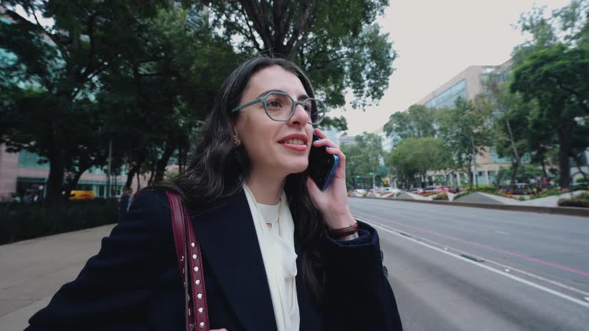 A professional young woman in glasses happily talks on her phone while standing on a busy city street