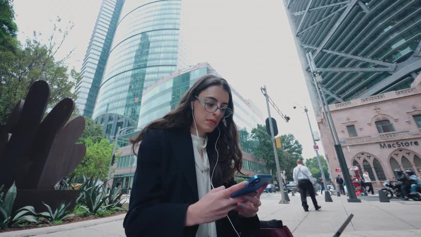 A young businesswoman with glasses and earphones works on her smartphone while sitting on a busy city street surrounded by towering skyscrapers