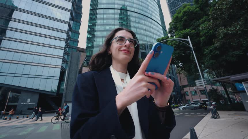 A stylish businesswoman with glasses smiles while using her smartphone in a modern city with towering glass skyscrapers
