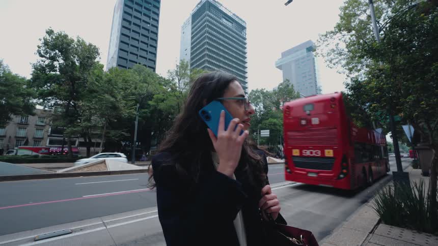A businesswoman makes a call on her smartphone while standing on a busy city street as a red bus drives past