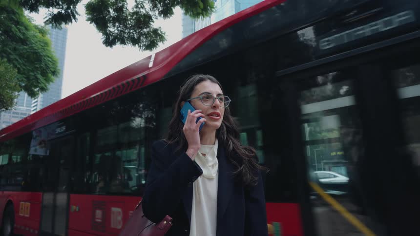A young professional woman stands on a bustling city street talking on her phone as a red bus pulls up behind her