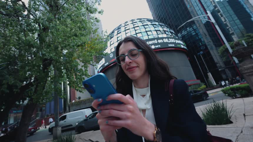 A confident businesswoman smiles while checking her smartphone with the Mexican Stock Exchange building in the background