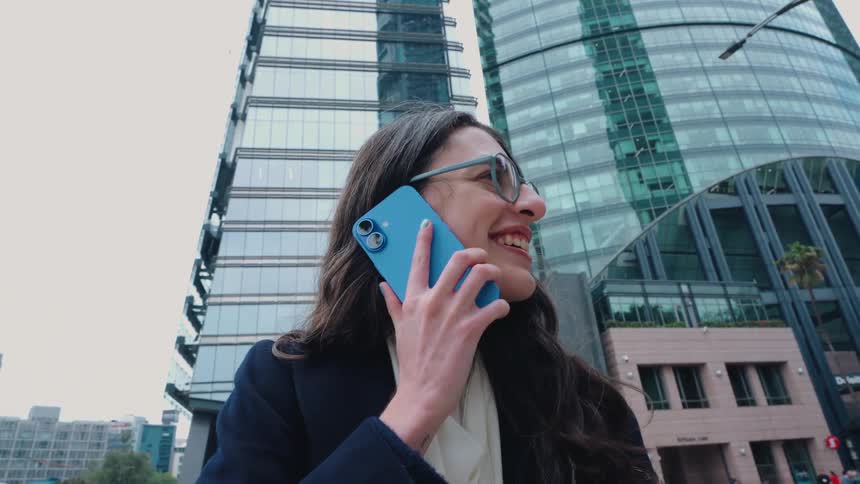 A happy businesswoman smiles while talking on her smartphone in front of a modern glass skyscraper in the city