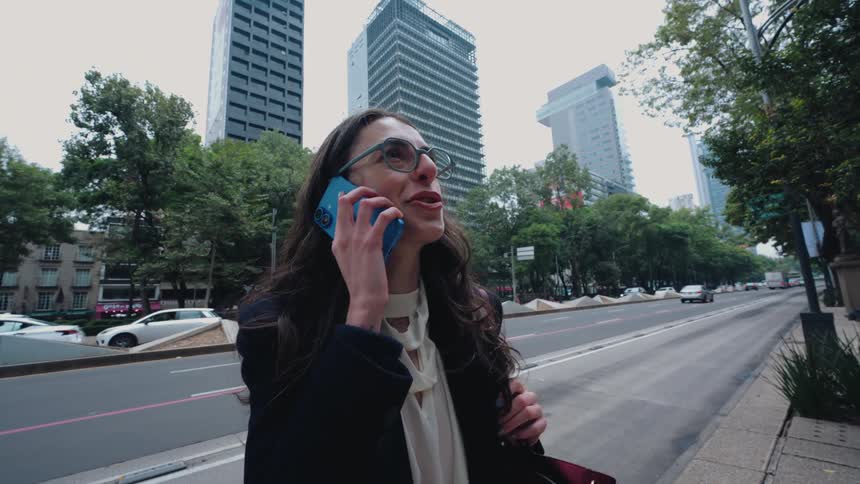 A happy businesswoman answers a call on her smartphone while standing on a busy city street with skyscrapers in the background