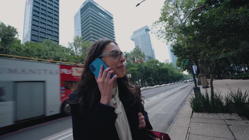 A stylish businesswoman smiles while talking on her smartphone on a busy city street with modern buildings and a tour bus driving by