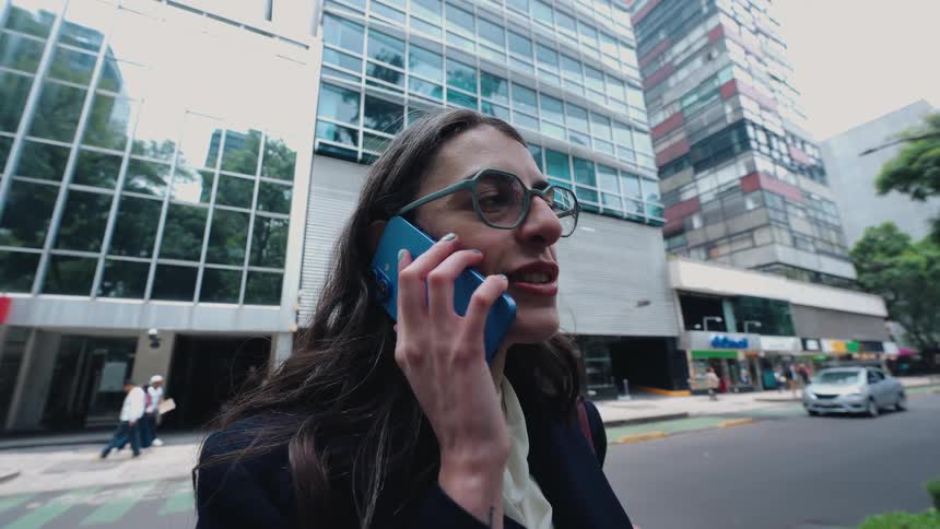 A businesswoman talks on her smartphone while standing on a busy city street with tall office buildings in the background