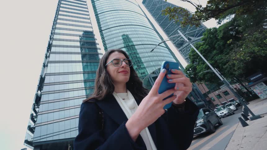 A stylish businesswoman smiles as she uses her smartphone on a busy city street with towering modern skyscrapers in the background