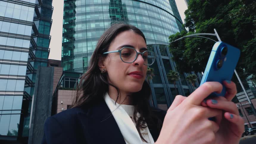A professional woman in glasses uses her smartphone while standing in front of a modern glass office building in the city