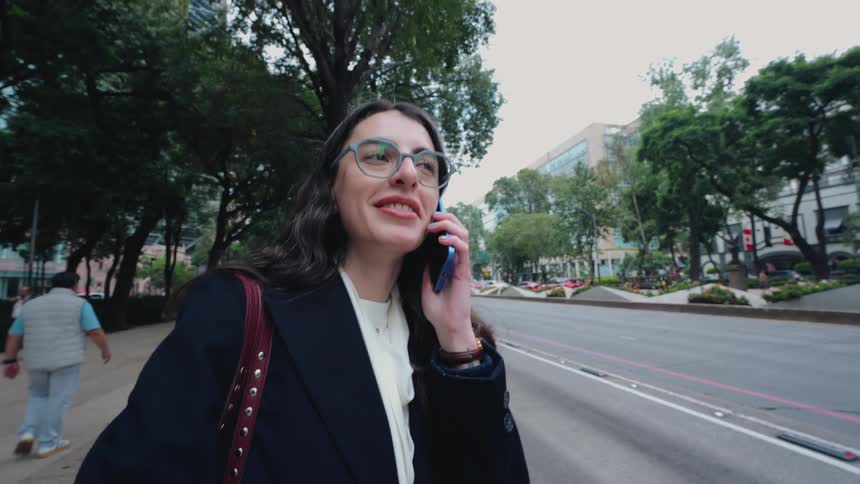 A stylish professional woman with glasses answers a phone call and smiles while talking on a city street