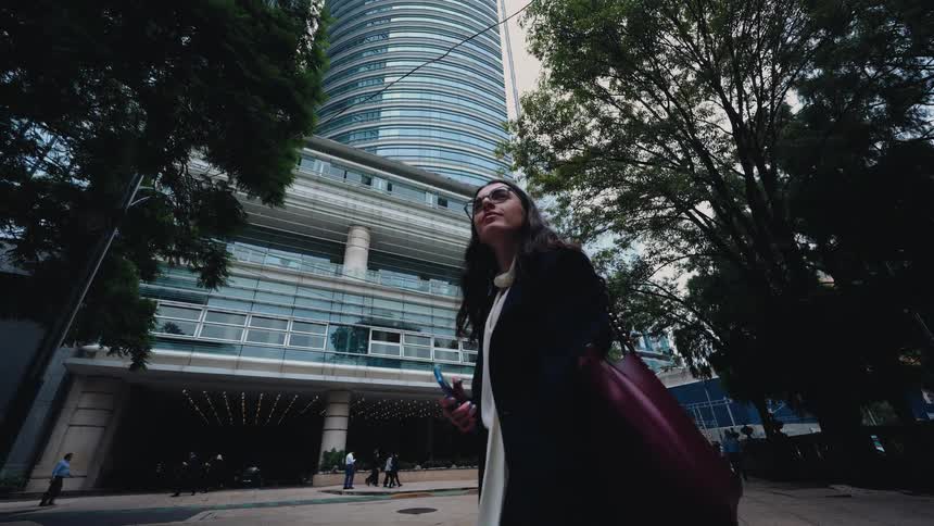 A professional woman walks in slow motion past a towering modern skyscraper in a bustling city financial district