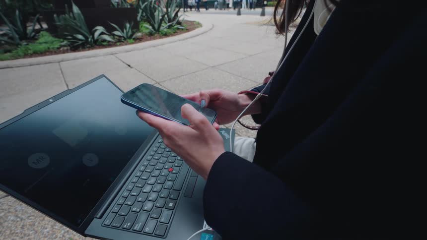 Close-up of a businesswoman's hands scrolling on her smartphone while attending a video conference on her laptop in an urban park